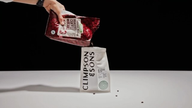 Coffee beans being poured from one red bag of roasted coffee beans into another white bag on a white table against a black background.