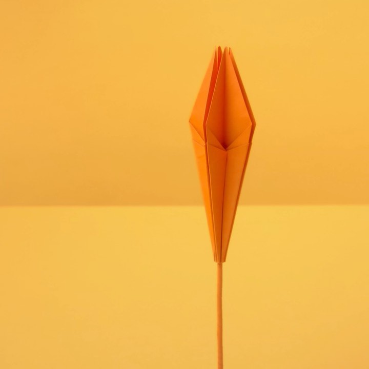 Orange paper origami flower on a light orange background. The paper flower is on a wood stem and stands upright.