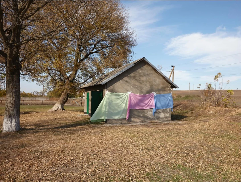 Exterior daytime shot of a small concrete building with clotheslines and laundry, next to a tree in a grassy, rural setting.