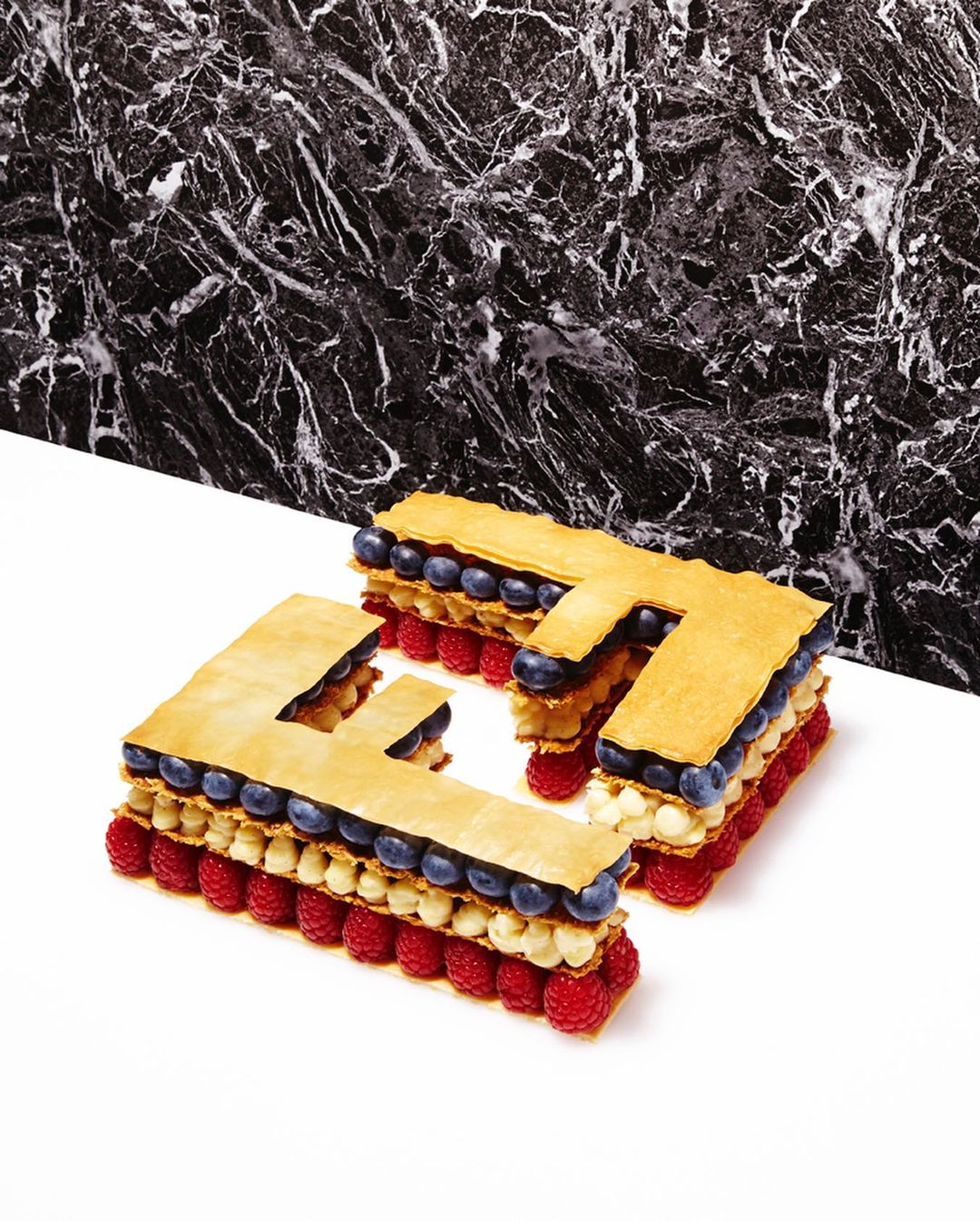 Geometric food art: Three 'F' shaped pastry desserts with raspberries and blueberries, arranged on a white surface against a black marble backdrop.