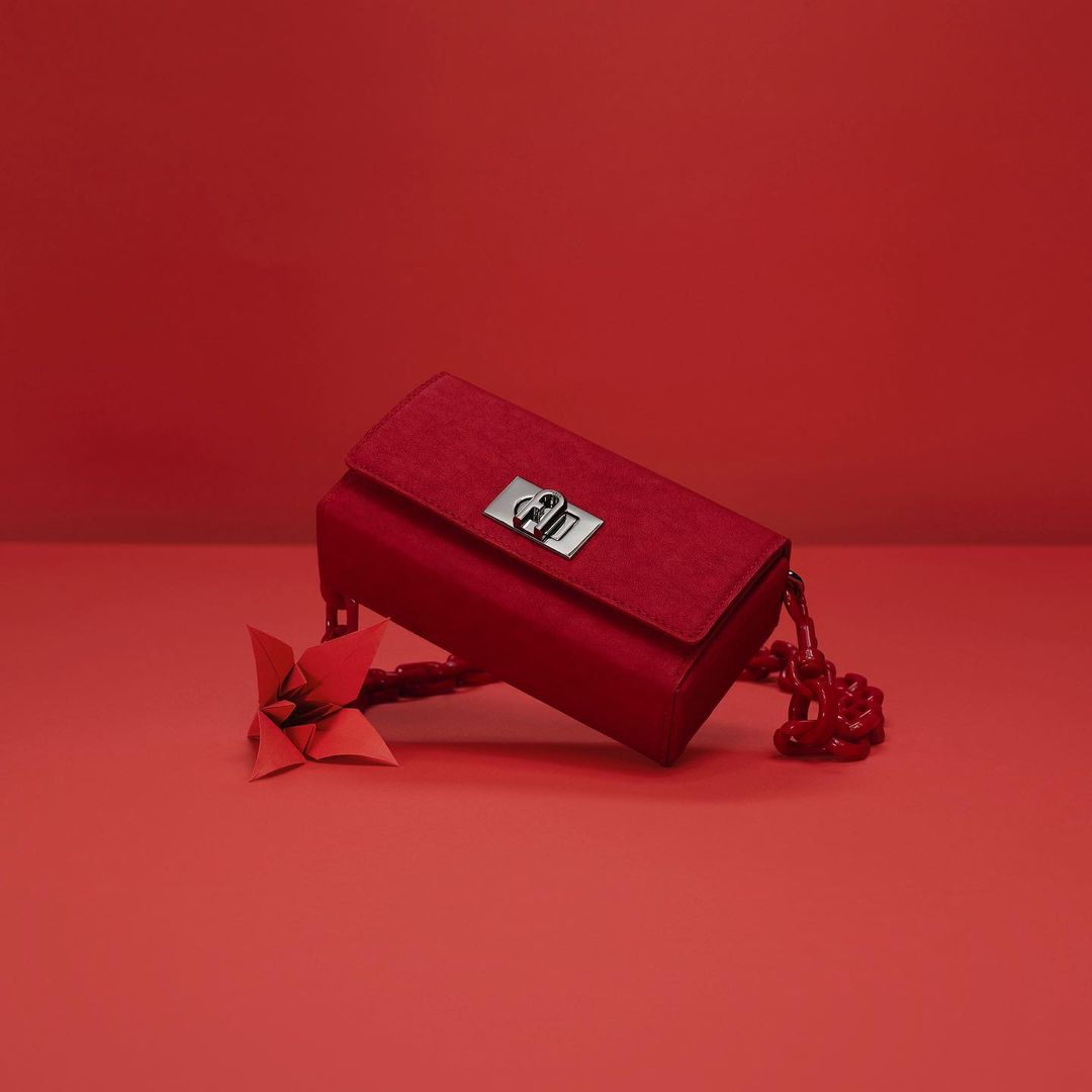 A red purse with a silver clasp and red chain handle lays on a red surface, next to a red origami flower, against a red background.