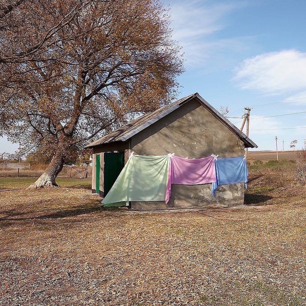Exterior view of a small, weathered building with colorful cloths drying outside on a sunny day, next to a large tree.