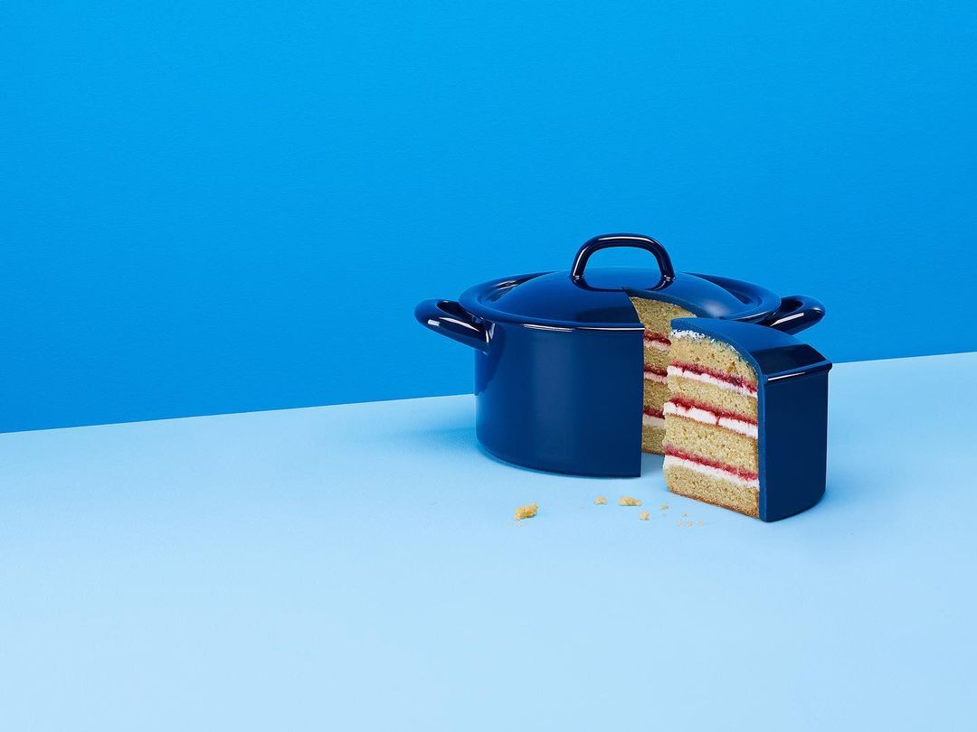 Blue enamel pot with a slice of multi-layered cake in front, against a split blue background. Conceptual food photography.