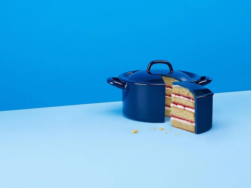 Blue enamel pot with a slice of multi-layered cake in front, against a split blue background. Conceptual food photography.