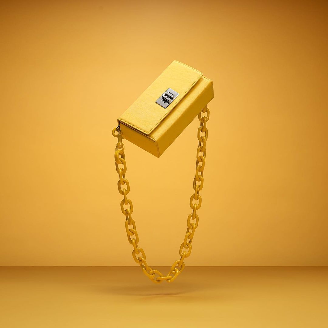 Yellow leather clutch with a chain handle floating against a matching gradient background in a fashion product shot for e-commerce.