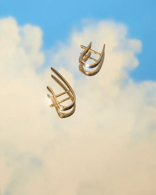 Studio shot of two gold earrings with a mirrored reflection against a blue sky and white clouds background for a fashion advertisement.