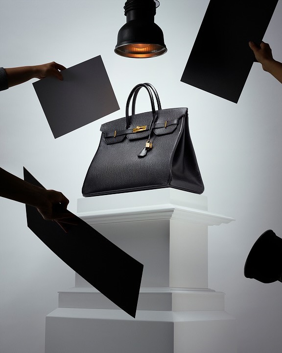Elevated view of a black leather Birkin bag with gold hardware on a white pedestal, surrounded by black and gray reflectors and studio lighting.