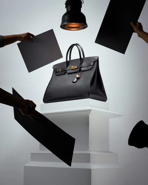 Elevated view of a black leather Birkin bag with gold hardware on a white pedestal, surrounded by black and gray reflectors and studio lighting.