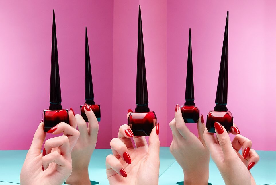Multiple bottles of red nail polish are displayed and held by hands with red nails, against a pink and teal background.