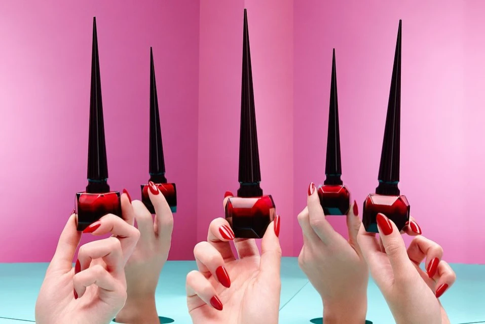 Multiple bottles of red nail polish are displayed and held by hands with red nails, against a pink and teal background.