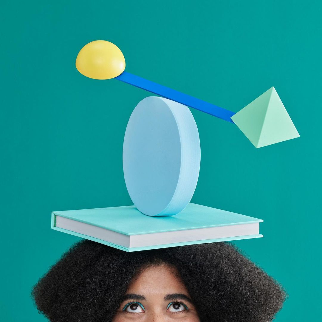 Woman balances a book with colorful geometric shapes on her head against a teal background in a studio shot using creative still life techniques.