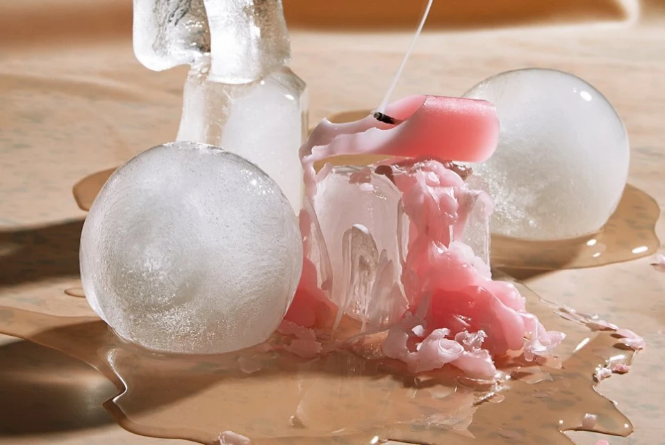 Still life of melting pink candle on clear ice, creating a visually striking contrast of warm and cold, on a light brown surface.