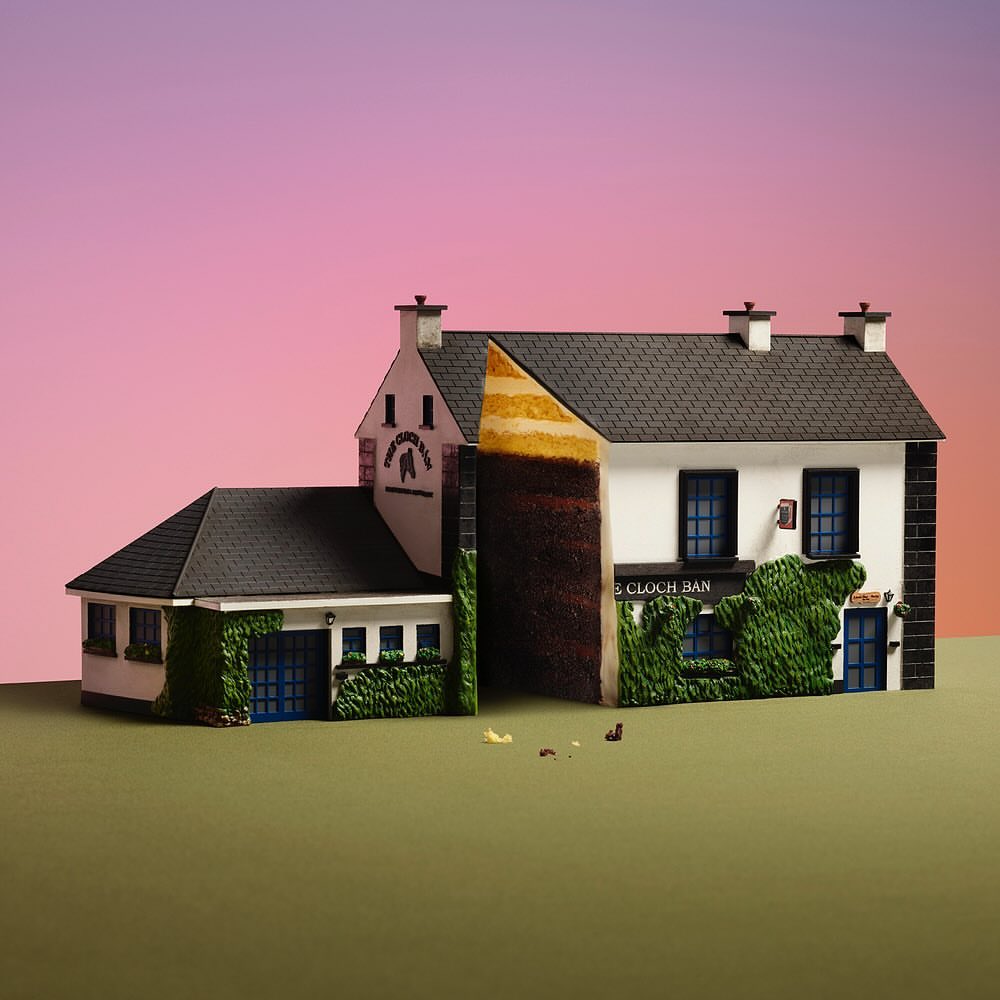 Architectural model of a two-story white building with black shingled roof, green vine accents, and logo on a pink gradient backdrop.