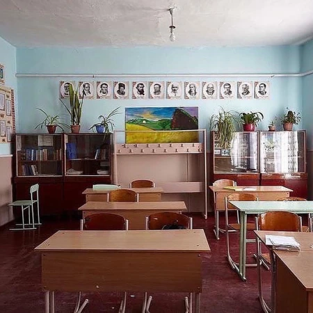 Empty classroom with wooden student desks, pale blue walls, dark red floor, and a row of framed portraits above.