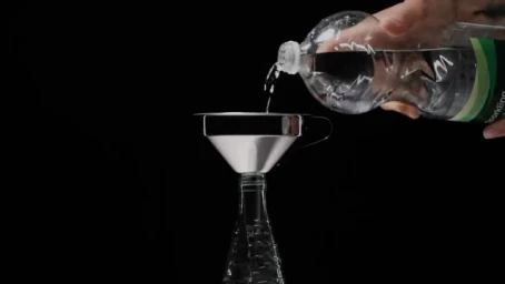 Water pouring from a plastic bottle through a metal funnel into another bottle against a black background.