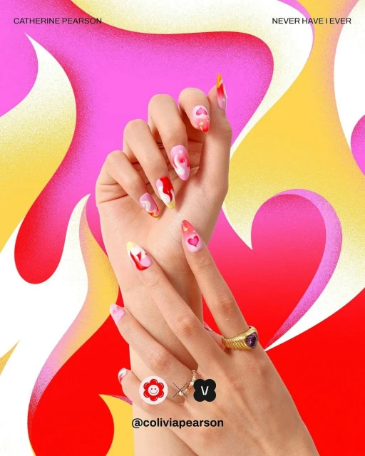 Close-up of hands with unique nail art featuring hearts and abstract designs, rings, and a vibrant pink, yellow, and red background.