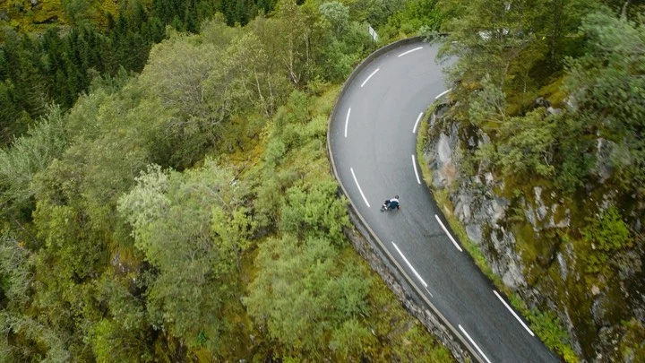 Aerial view of a person skateboarding down a winding road through a lush green forest with mossy rocks and a sharp curve.