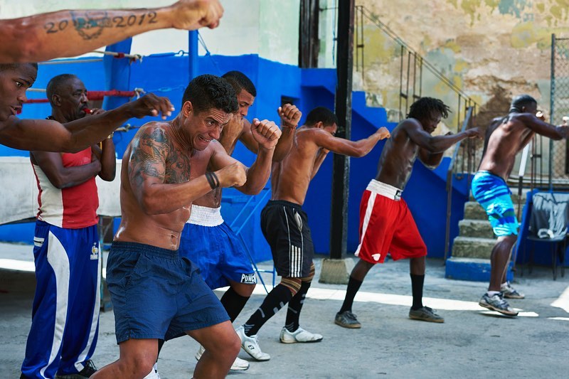 Several muscular, shirtless men with boxing shorts and hand wraps throwing punches in a boxing gym with natural light, Cuba.