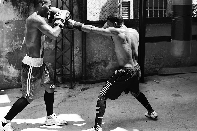 Two shirtless adult males in boxing gear exchanging punches in an outdoor boxing ring, photographed in black and white.