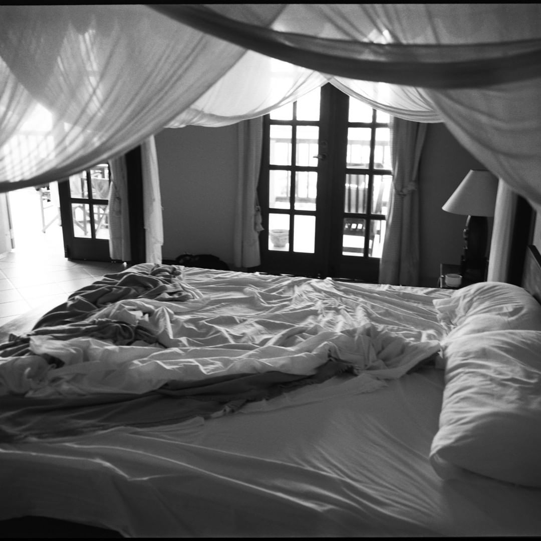 Black and white shot of a bedroom with a large bed covered in white linens and pillows, featuring windows and a draped canopy.