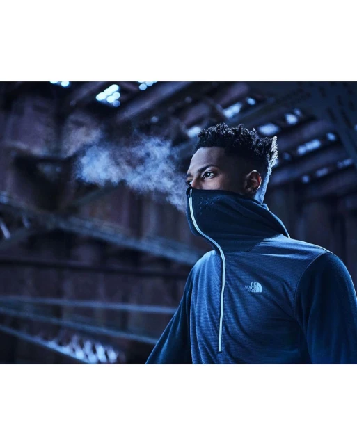 Close-up of a man wearing a navy blue, zip-up hooded athletic jacket with a close-fitting hood, breath visible in the cool air, dim industrial backdrop.