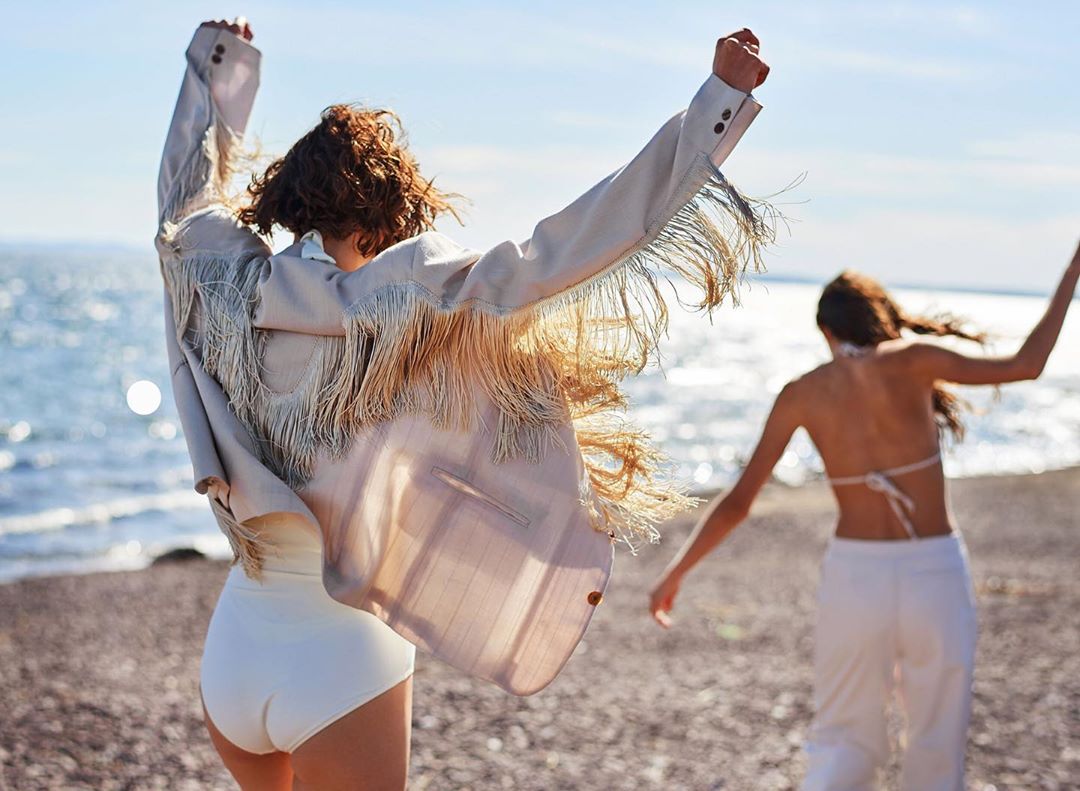 Two women in fashionable outfits are gleefully running on a beach with arms raised, under the bright daylight and sunny sky, showcasing an open-air lifestyle.