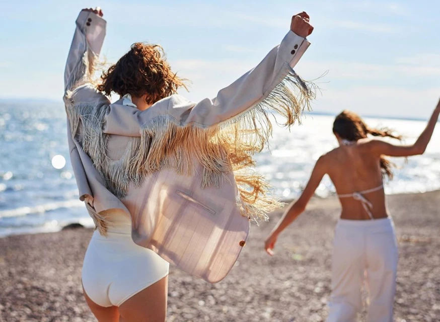 Two women in fashionable outfits are gleefully running on a beach with arms raised, under the bright daylight and sunny sky, showcasing an open-air lifestyle.