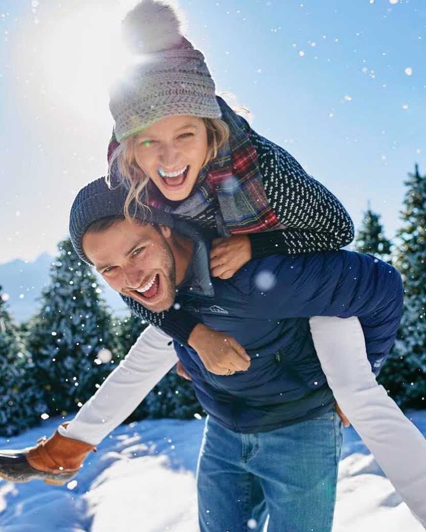 A happy couple enjoys a winter day in a snowy forest. A smiling woman rides piggyback on a smiling man, with snow falling around them.