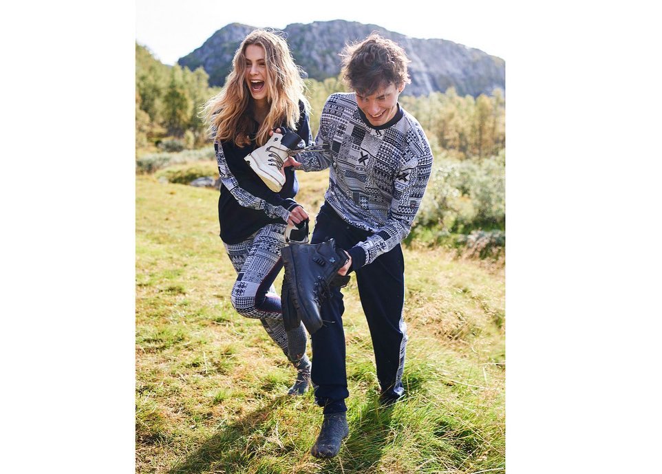 Two people in patterned sweaters hold hiking boots in a grassy, mountainous outdoor setting on a bright day. Fashion shoot.