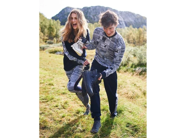 Two people in patterned sweaters hold hiking boots in a grassy, mountainous outdoor setting on a bright day. Fashion shoot.
