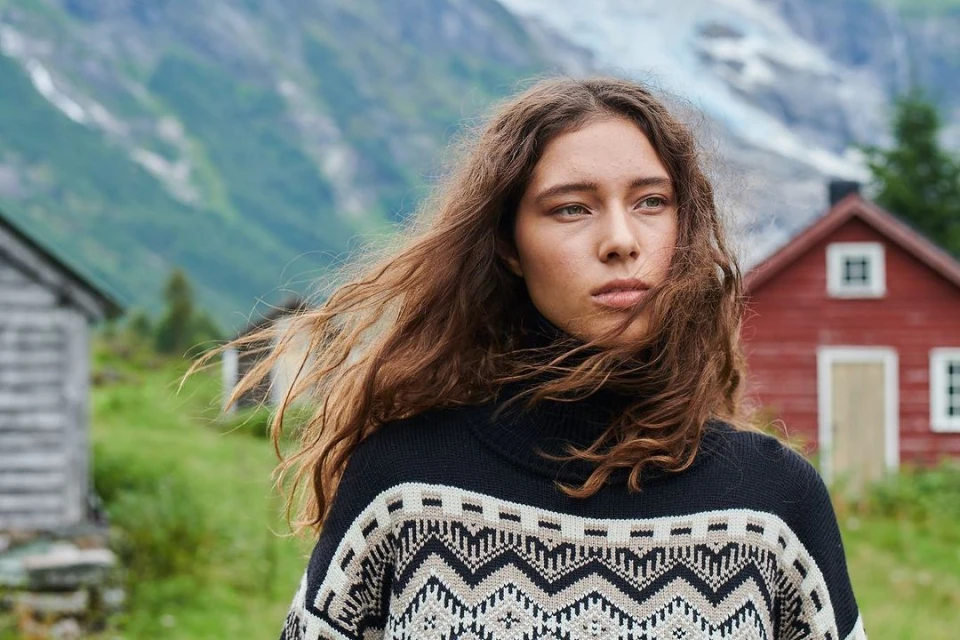Adult female in a black and beige patterned sweater posing outdoors with a mountain and small buildings in the blurred background.