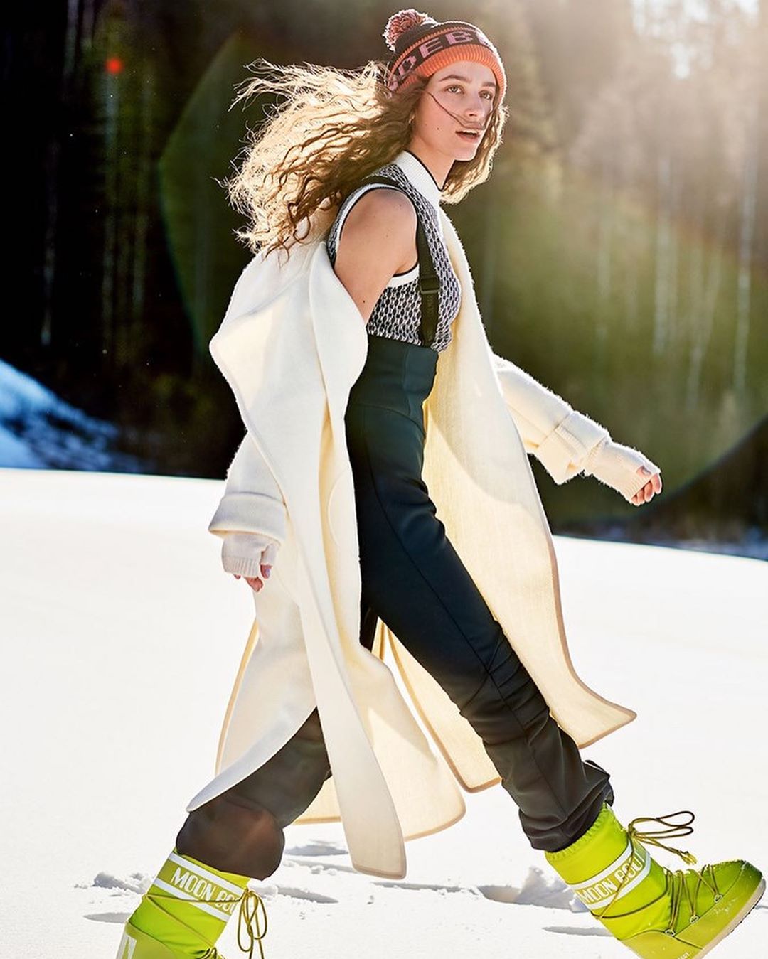 A fashionable young woman confidently walks through a snowy landscape wearing bright snow boots, a long coat, and a beanie.