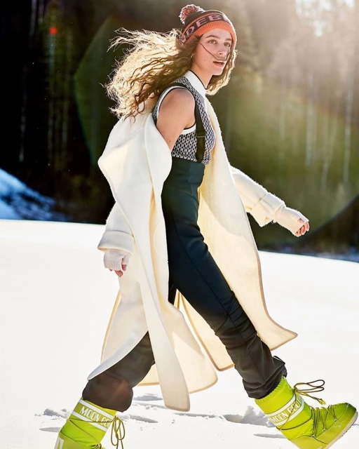 A fashionable young woman confidently walks through a snowy landscape wearing bright snow boots, a long coat, and a beanie.