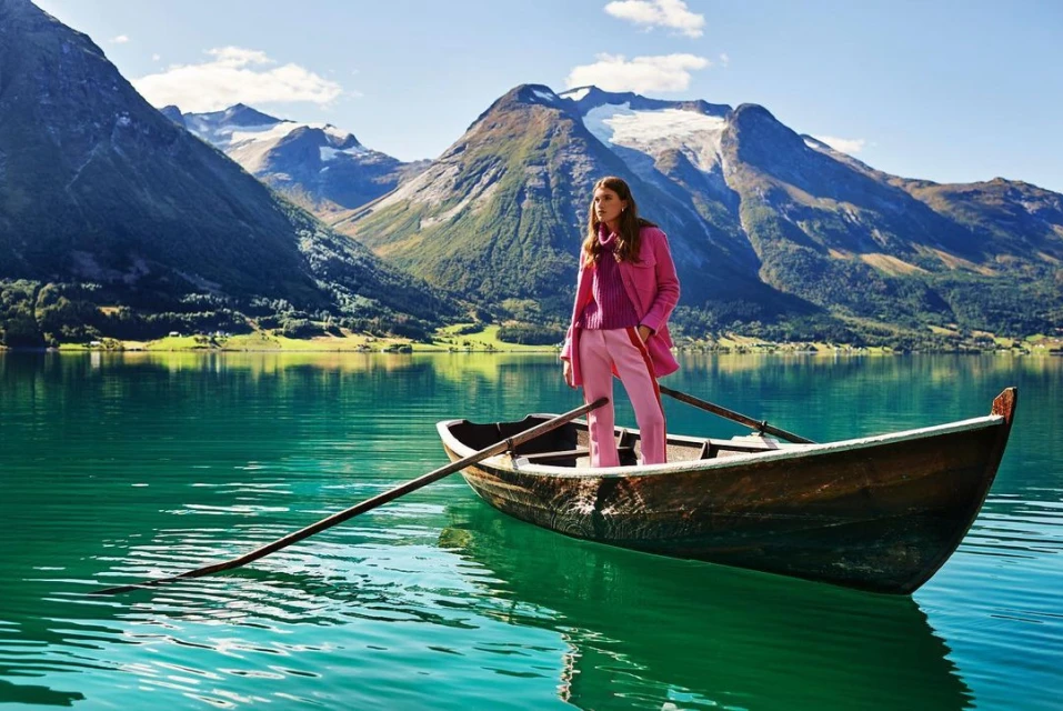 Woman in pink suit standing in rowboat on calm lake with snow-capped mountains in the distance, captured on a sunny day outdoors.