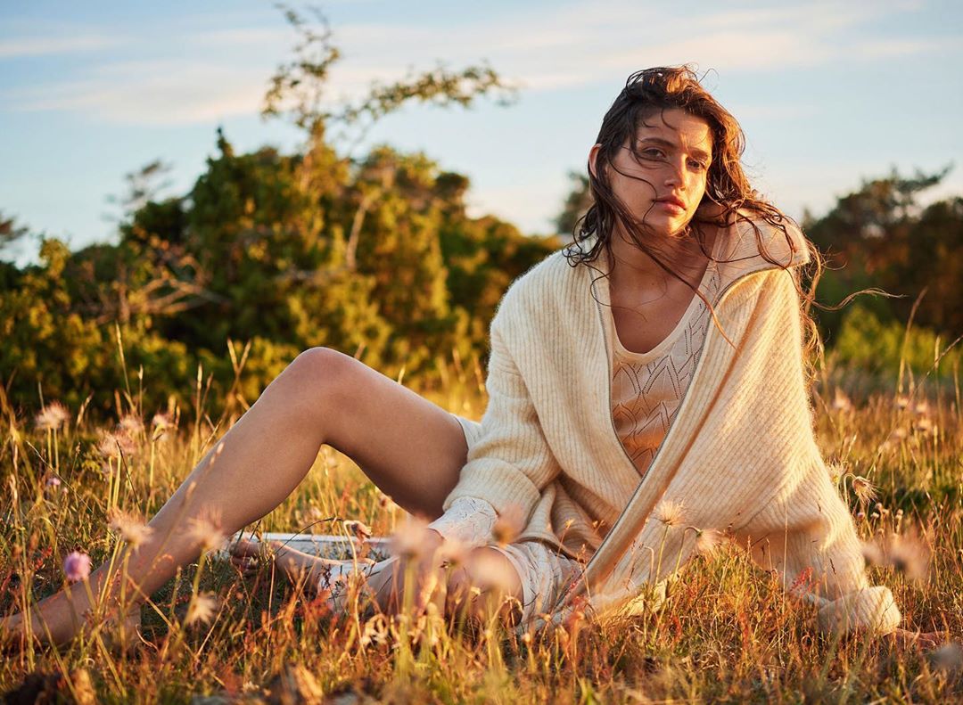 A woman with long brown hair is sitting in a field of tall grass and wildflowers, wearing a sweater and a knitted top and shorts.