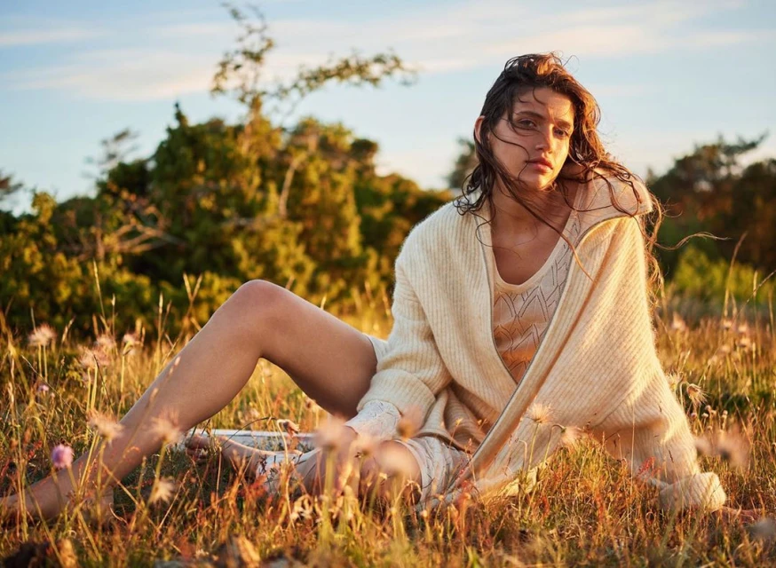 A woman with long brown hair is sitting in a field of tall grass and wildflowers, wearing a sweater and a knitted top and shorts.