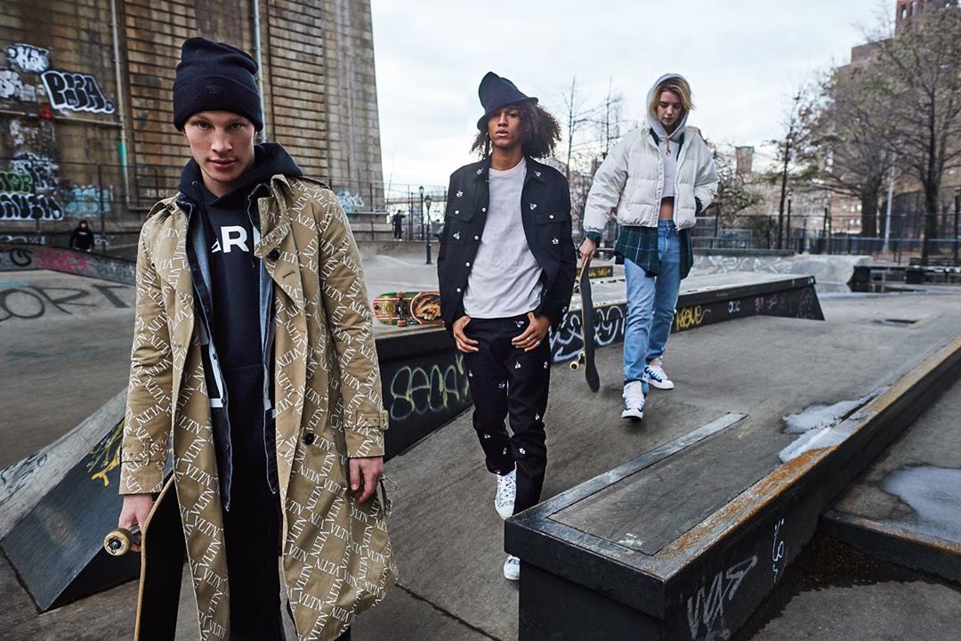 Three stylish young adults walk in a concrete skatepark with a bridge in the background wearing fashionable streetwear.