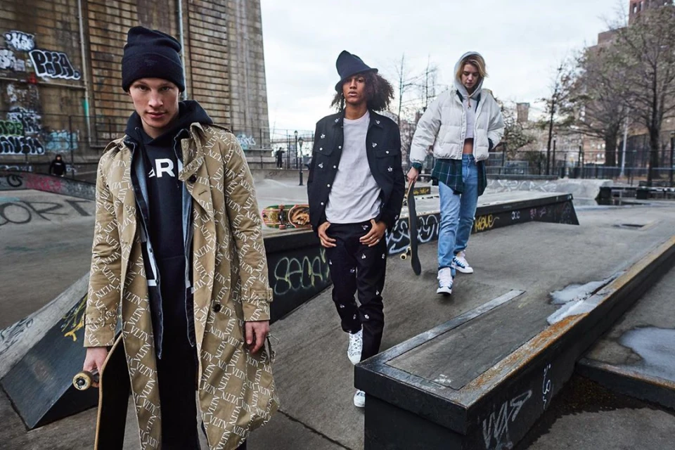 Three stylish young adults walk in a concrete skatepark with a bridge in the background wearing fashionable streetwear.