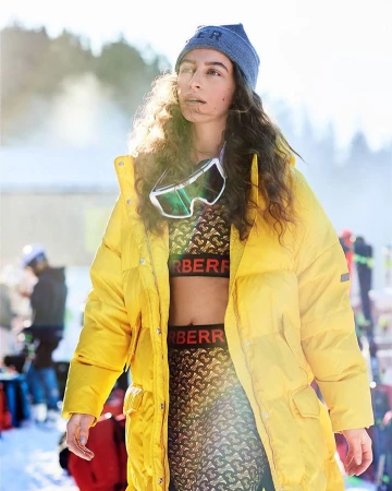 Woman in a yellow puffer coat and knit hat, accessorized with ski goggles, posing outdoors in a snowy, mountainous environment.