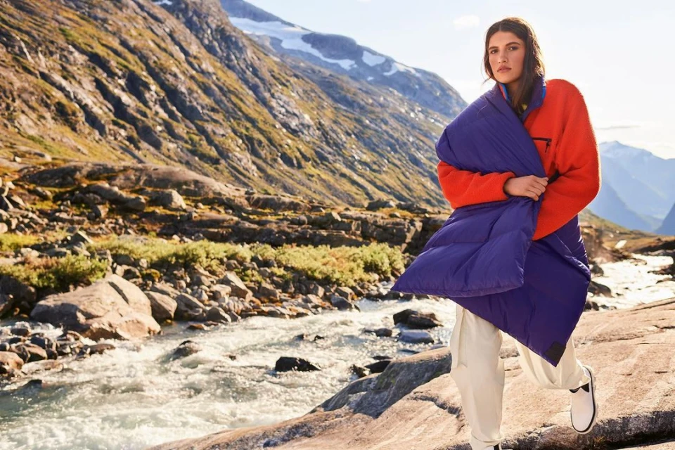A woman stands on rocks by a river in a mountain landscape, wrapped in a blue quilted blanket and wearing a red fleece and cream pants.
