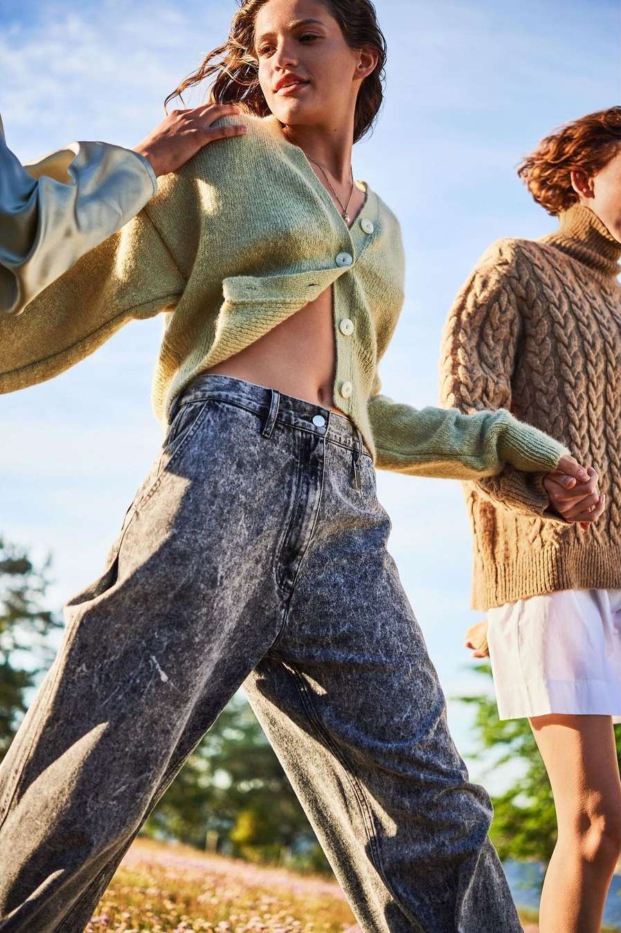 Three women model casual spring fashion outdoors, featuring cardigans and wide-leg denim. Sunlight illuminates the meadow setting.