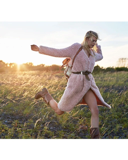 Woman in a coat runs through tall grass at sunset, wearing boots and backpack in an outdoor nature setting.