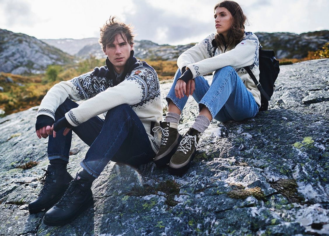 Two people sit on a rock formation in a mountain landscape, wearing patterned sweaters, jeans, and hiking boots; cloudy sky in background.