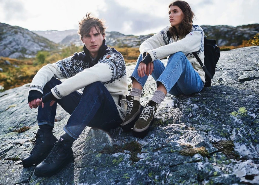 Two people sit on a rock formation in a mountain landscape, wearing patterned sweaters, jeans, and hiking boots; cloudy sky in background.