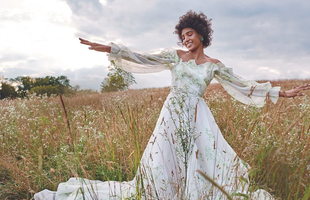 A woman in a white floral dress stands in a field of wildflowers, smiling and holding her arms out with cloudy sky in the background.