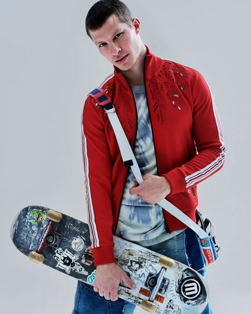 Adult male poses in a red jacket with a skateboard and shoulder bag in a photo studio setting with soft lighting and a neutral background.