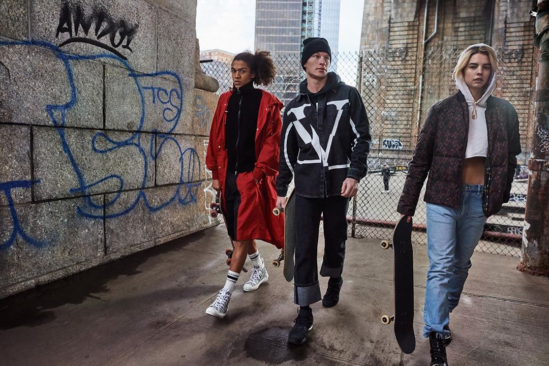 Three models walk on a city street with skateboards, wearing jackets and jeans, captured in a medium shot with urban graffiti.