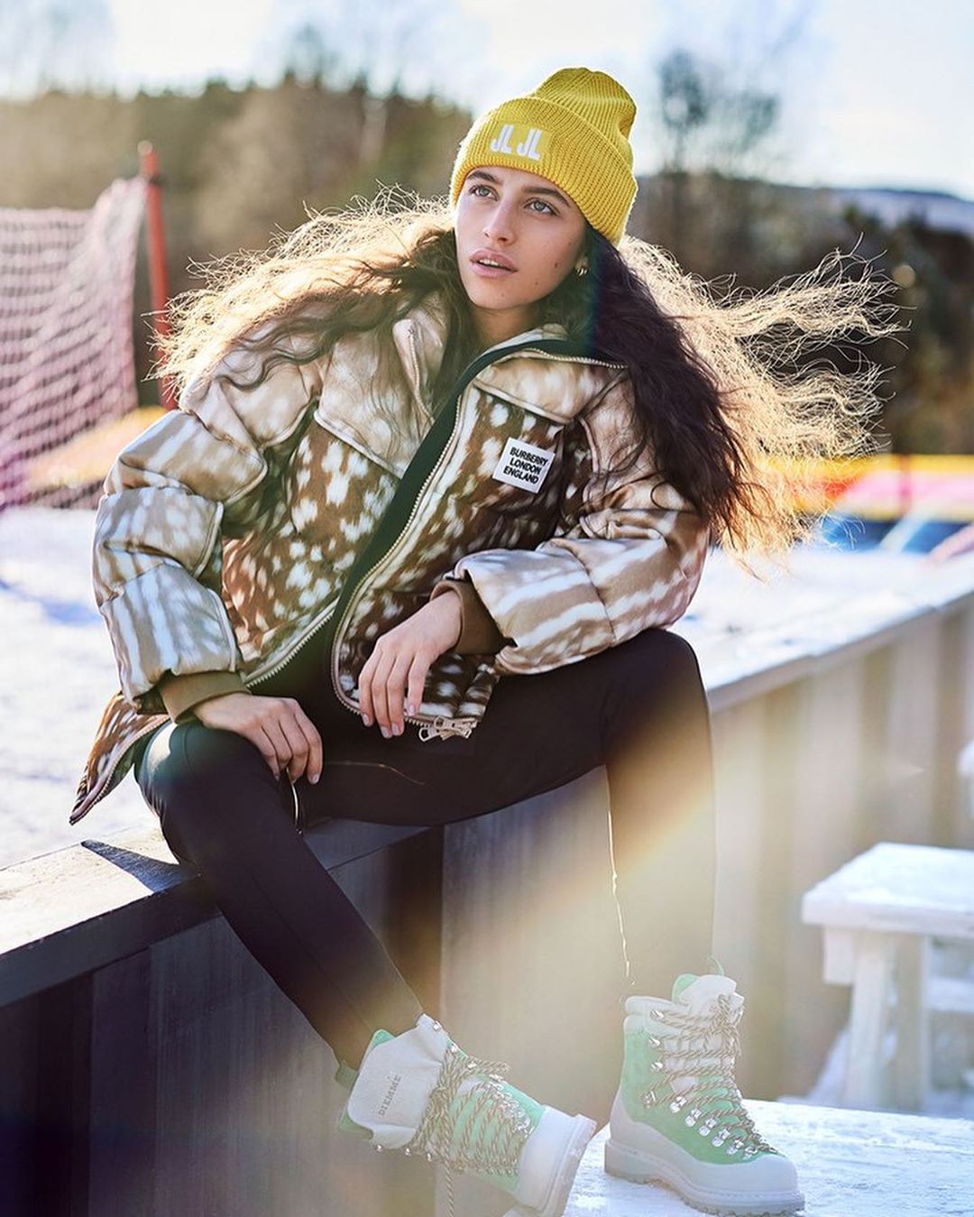 Woman in patterned puffer jacket and yellow beanie sitting outside on a bench, wearing decorative boots, posing in a medium shot.