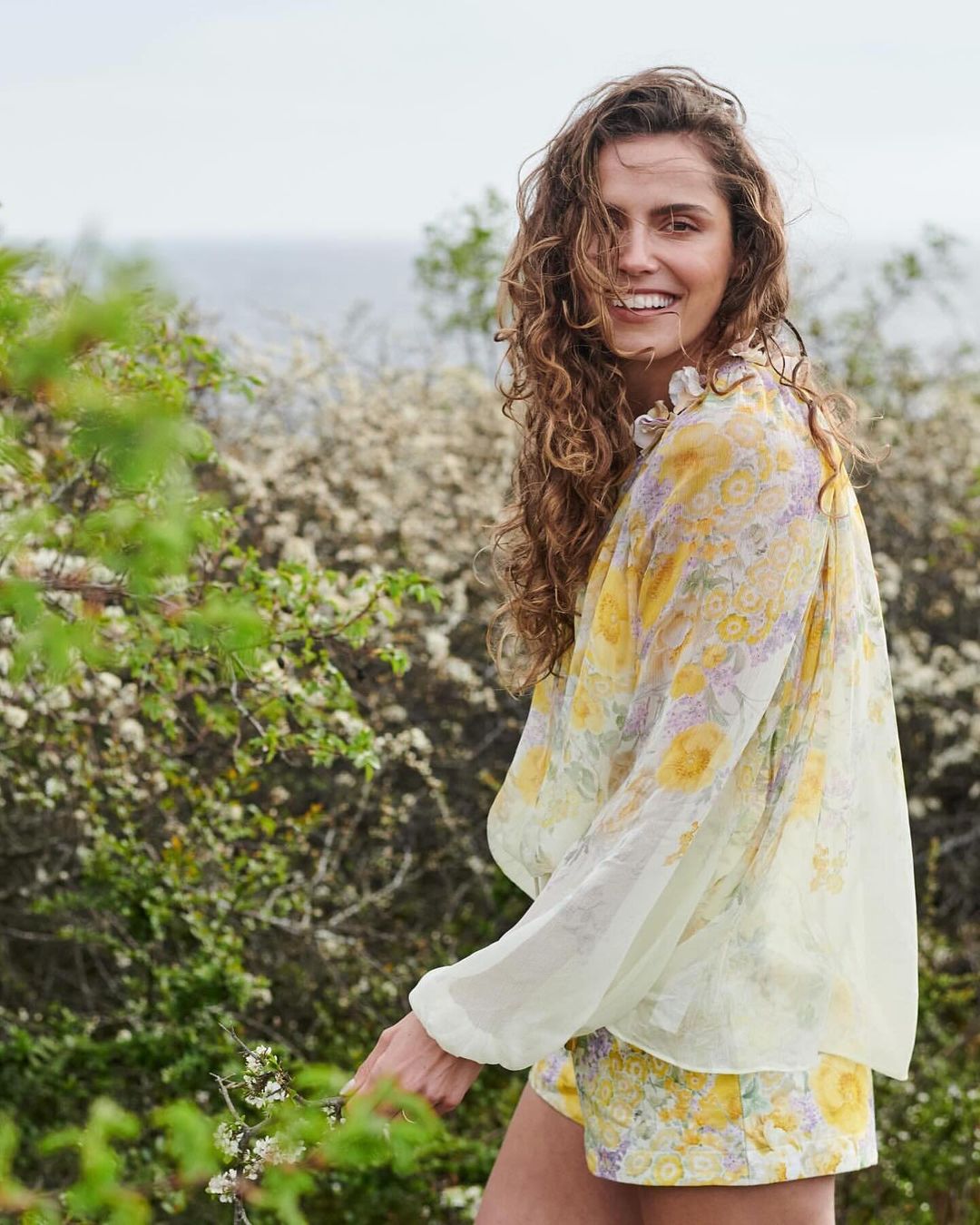 A smiling woman with curly hair is posing outdoors in a floral blouse and shorts, standing in a natural setting with greenery.