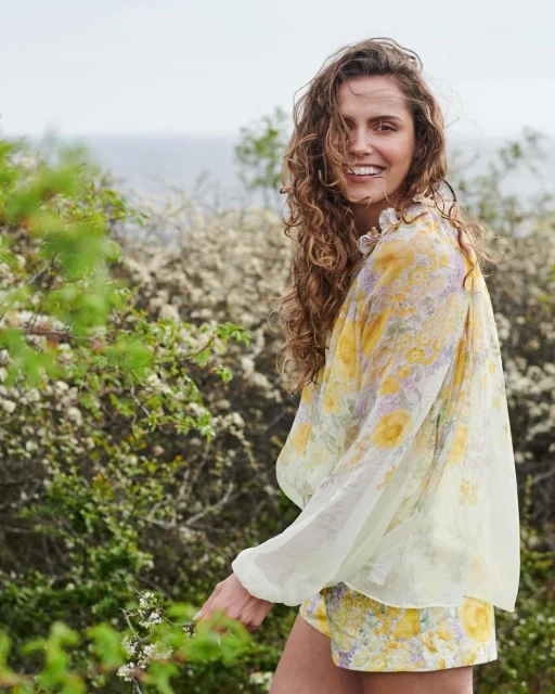 A smiling woman with curly hair is posing outdoors in a floral blouse and shorts, standing in a natural setting with greenery.